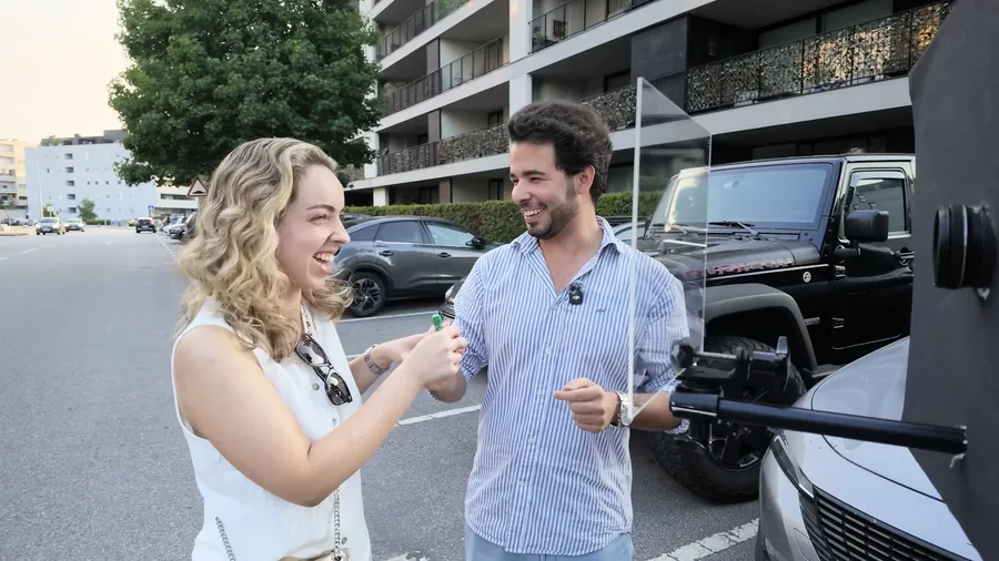 Duas pessoas a rir num parque de estacionamento durante uma rodagem, uma delas a segurar um marcador e uma placa de acrílico